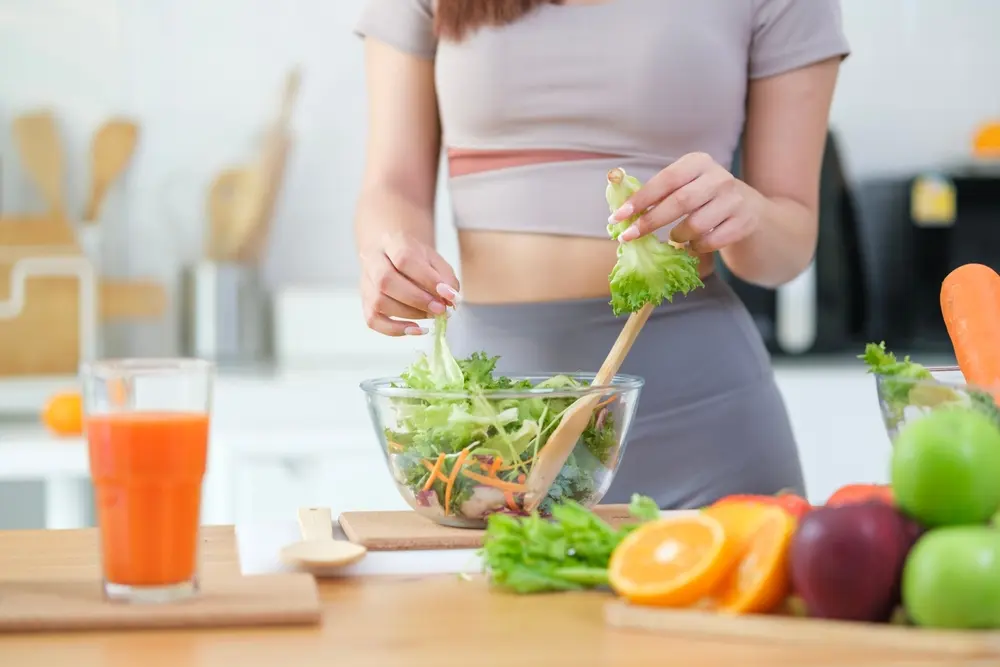 Person in a gray outfit tossing a green salad in a glass bowl on a bright kitchen counter. Fresh veggies and juice are nearby.
